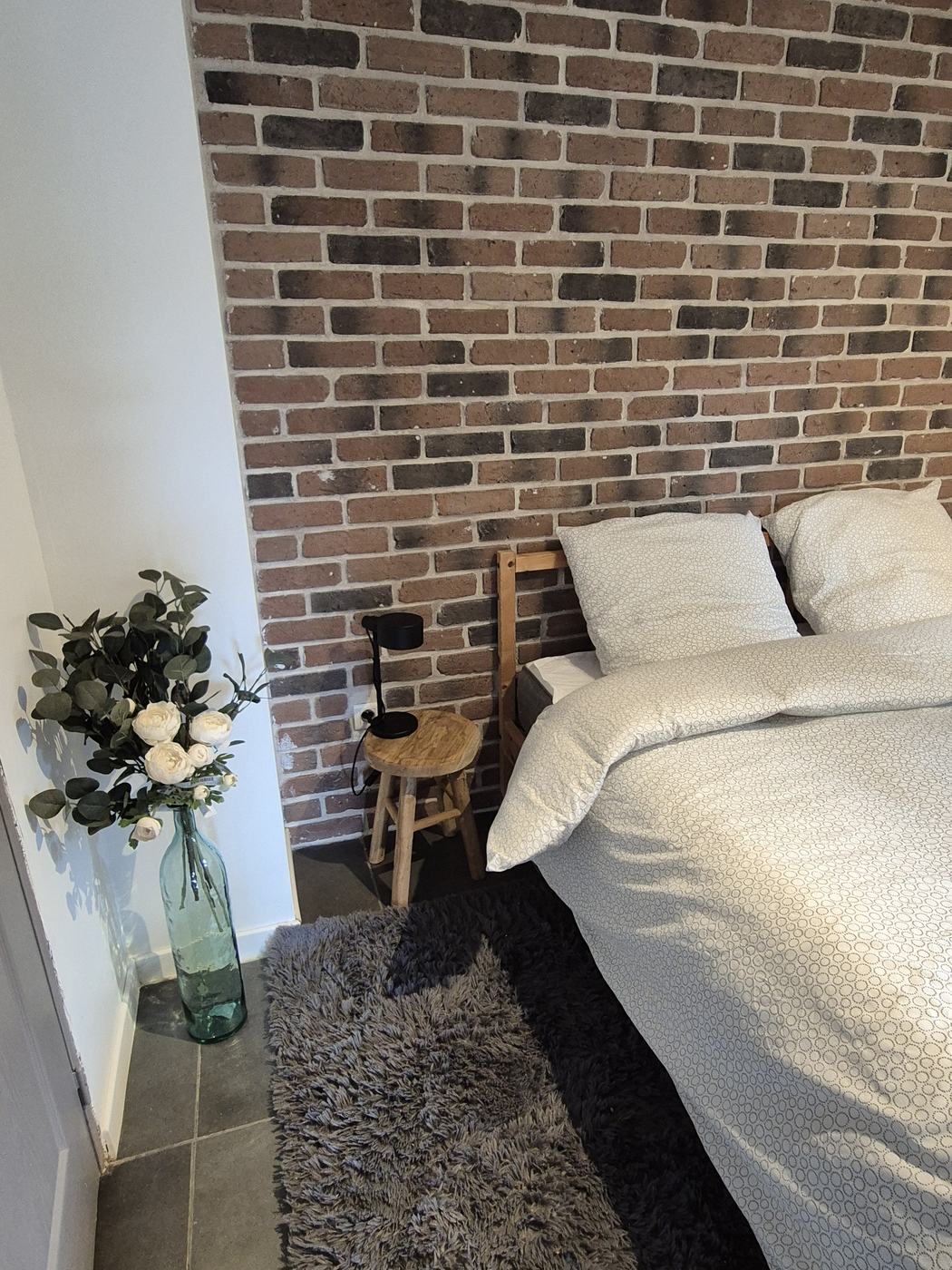 Détail de la chambre : tabouret en bois, lampe noire et linge de lit clair